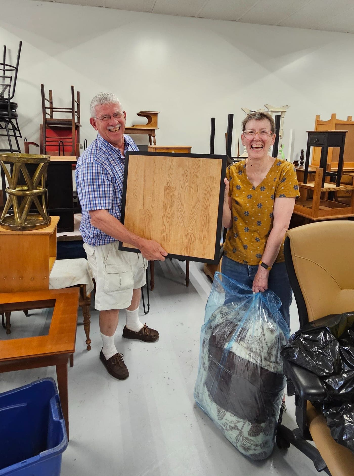 Two smiling HomeAhead volunteers in the furniture storage room, one holding a wooden tabletop and one a bag of bedding