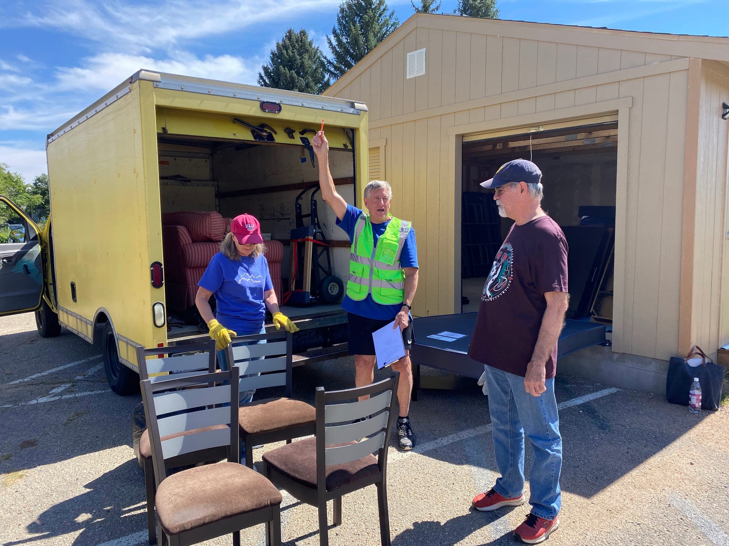Three HomeAhead volunteers with four donated dining chairs beside an open yellow box truck, one in a safety vest holding a clipboard